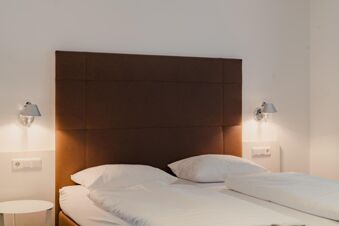 Double bed with white linens and pillows, brown padded headboard, two wall-mounted lamps, and side table at Gasthaus Backmulde - Hotel