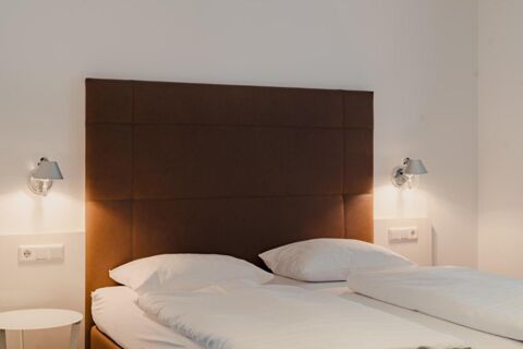 Double bed with white linens and pillows, brown padded headboard, two wall-mounted lamps, and side table at Gasthaus Backmulde - Hotel