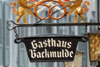 Tanda penginapan bersejarah Gasthaus Backmulde - Hotel dengan dua singa emas dan pretzel panggang sebagai hiasan di depan jendela.