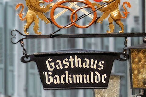 Tanda penginapan bersejarah Gasthaus Backmulde - Hotel dengan dua singa emas dan pretzel panggang sebagai hiasan di depan jendela.
