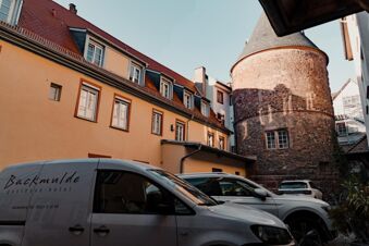 Several parked cars and a van with Backmulde gasthaus hotel logo are next to a yellow building and a round stone tower at Gasthaus Backmulde - Hotel