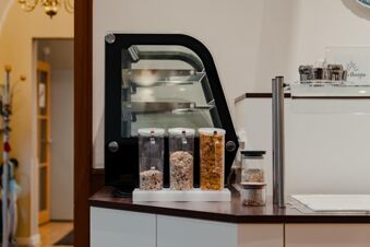 Three transparent cereal dispensers with different cereals on a counter next to a black bread slicer at Gasthaus Backmulde - Hotel