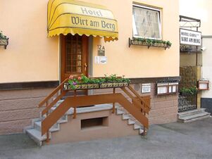 Eingangsbereich und Fassade von Hotel Wirt am Berg mit gelbem Sonnendach, Blumenk&auml;sten und mehreren Treppen zum Eingang.