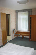 Helles Hotelzimmer im Hotel Wirt am Berg mit floral gemusterter Tapete, Holzstuhl, Gardinen und offener Badezimmert&uuml;r.