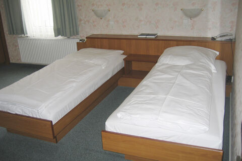 Zweibettzimmer im Hotel Wirt am Berg mit zwei Einzelbetten, wei&szlig;er Bettw&auml;sche, blumenverziertem Tapetenmuster und Fenster mit Vorh&auml;ngen.