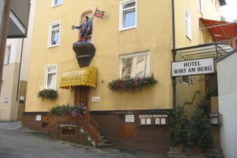 Die gelbe Fassade des Hotel Wirt am Berg mit Blumenk&auml;sten, einem Dachvorsprung und einem historischen Figurenbanner &uuml;ber dem Eingang.