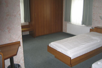 Helles, einfach eingerichtetes Zimmer mit Holzm&ouml;beln und Bett im Hotel Wirt am Berg mit blumiger Tapete und Vorh&auml;ngen.