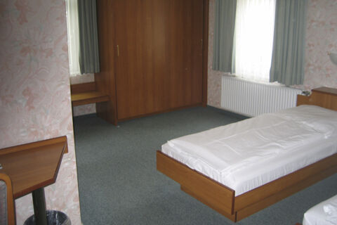 Helles, einfach eingerichtetes Zimmer mit Holzm&ouml;beln und Bett im Hotel Wirt am Berg mit blumiger Tapete und Vorh&auml;ngen.
