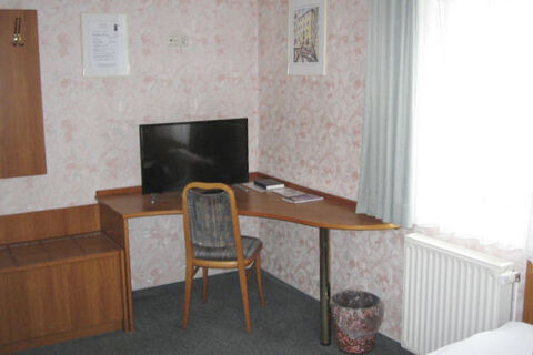 Kleiner Arbeitsbereich mit Fernseher, Holzstuhl und Fenster mit Gardine in einem Zimmer des Hotel Wirt am Berg.