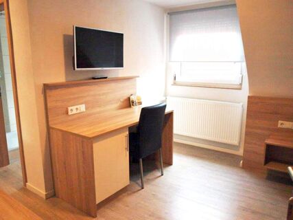 Minimalist room in Gasthof zur Sonne with a wooden desk, black chair, wall-mounted TV, radiator, and window with white blind