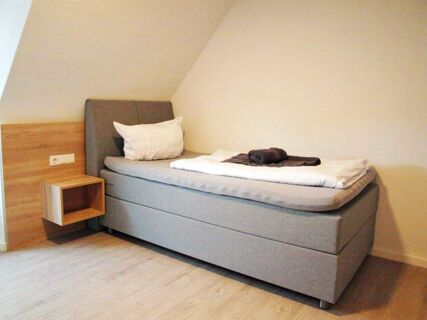 A single grey bed with a white pillow, folded white bedding, and dark towels beside a wooden nightstand in a Gasthof zur Sonne room