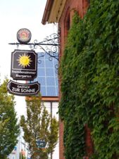 A decorative iron sign for Gasthof zur Sonne with a sun logo hangs from a brick building covered in green ivy leaves