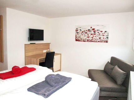 A clean hotel room at Gasthof zur Sonne with a double bed, folded towels, a desk with chair, wall-mounted TV, and a gray sofa with cushions