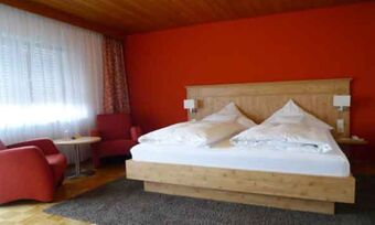 Svetla hotelska soba z rdečo steno, leseno posteljo in udobnimi fotelji v elegantno opremljenem hotelu Krone.