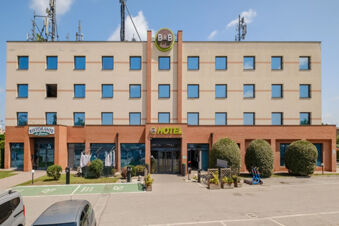 Facade of B&B Hotel Ferrara showing rectangular windows, a restaurant on the left, bushes, parked cars, and scooters outside