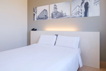 Minimalist hotel room with white bed linens, two pillows, white headboard, three black and white city photos, and a phone at B&B Hotel Ferrara