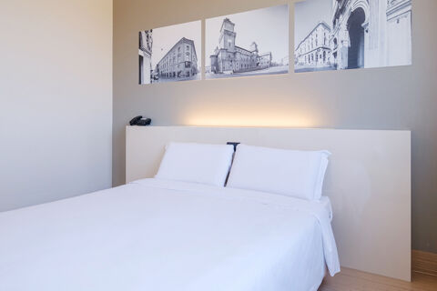 Minimalist hotel room with white bed linens, two pillows, white headboard, three black and white city photos, and a phone at B&B Hotel Ferrara