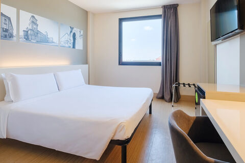 Minimalist hotel room at B&B Hotel Ferrara with a large bed, white linens, wooden floor, window, TV, and desk chair