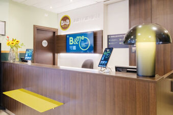 Reception desk area at B&B Hotel Ferrara with wooden counter, yellow bench, digital screens, flowers, and modern lamp