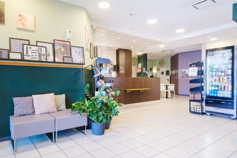 Waiting area with couches, cushions, potted plants, artwork, a reception desk, and a vending machine inside B&B Hotel Ferrara