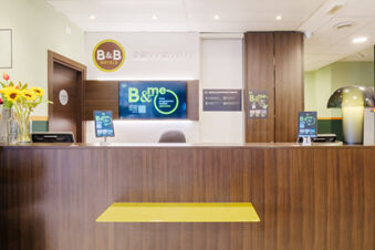 Reception desk with two digital screens, a sign reading B&B Hotels, flowers in a glass vase and a lamp at B&B Hotel Ferrara