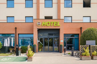 Entrance of B&B Hotel Ferrara with automatic glass doors, several windows, a green electric vehicle charging area and plants outside
