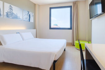 Bright hotel room at B&B Hotel Ferrara with a double bed, framed city photos, window, TV, and green chairs