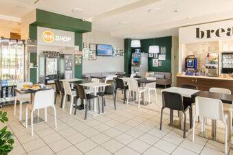 Spacious dining area with white and black chairs, tables, snack vending machines, and B&B Hotel Ferrara shop corner