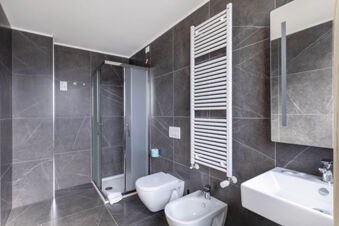 Modern bathroom with gray tiled walls, shower with frosted glass doors, toilet, bidet, and white sink at B&B Hotel Ferrara