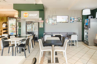 Dining area of B&B Hotel Ferrara with black and white chairs at tables, a fridge with drinks, and a wall-mounted TV