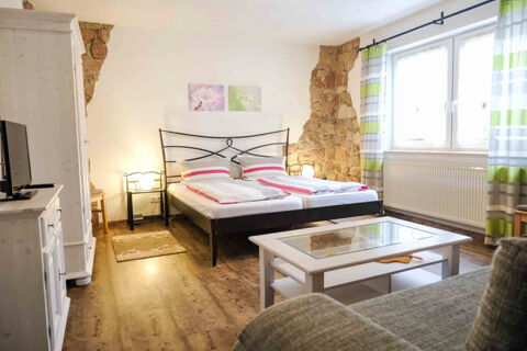 Double bed with striped bedding, bedside tables with lamps, a TV cabinet, and window with green-striped curtains in G&auml;stehaus Blank room