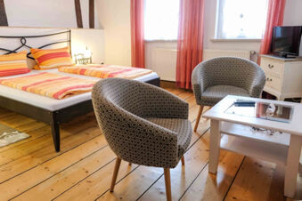 A G&auml;stehaus Blank room with two single beds with striped bedding, two patterned armchairs, a white table, and a TV on a small cabinet