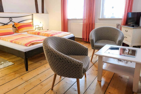 A G&auml;stehaus Blank room with two single beds with striped bedding, two patterned armchairs, a white table, and a TV on a small cabinet