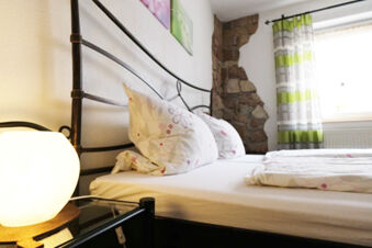 Bed with floral pillows, white and pink bedding, stone wall, green and white striped curtains, and a lit lamp in G&auml;stehaus Blank
