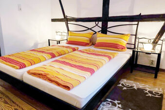 Double bed with yellow and orange striped bedsheets and pillows, black metal frame, two lamps on black side tables in G&auml;stehaus Blank bedroom