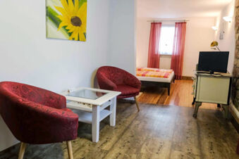 Two red patterned armchairs with a white glass top table between them and a bed with a colorful blanket in G&auml;stehaus Blank