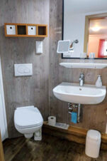 Compact bathroom in G&auml;stehaus Blank featuring a wall-mounted toilet, white sink with mirror, shelf with two cups, and wooden panel walls