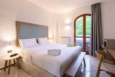 Hotel room at B&B HOTEL Ischia San Nicola featuring a double bed with white linens, wooden bench, side table, and glass door with gray curtains
