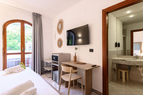 Hotel room at B&B HOTEL Ischia San Nicola with a wooden desk, wall-mounted TV, two round mirrors, and balcony doors with curtains