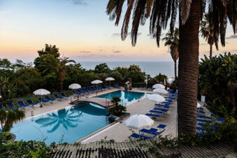 Outdoor swimming pool area with blue lounge chairs, white umbrellas, palm trees, and ocean view at B&B HOTEL Ischia San Nicola
