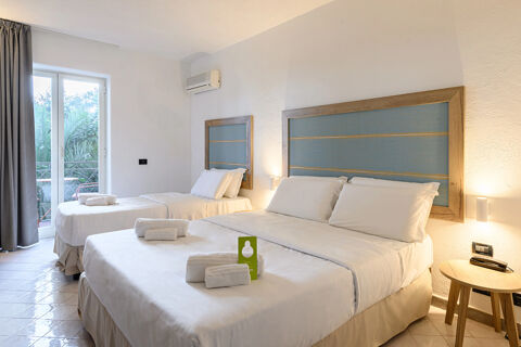 A clean and bright hotel room at B&B HOTEL Ischia San Nicola with two beds, white linens, towels, a small wooden table, and a window with gray curtains showing greenery outside