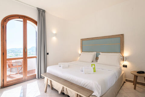 Bright hotel room at B&B HOTEL Ischia San Nicola with a large bed, wooden bench, arched door, and balcony seating outdoors