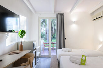 Hotel room at B&B HOTEL Ischia San Nicola with two single beds, bedside bench, desk with chair, TV, and glass doors to balcony