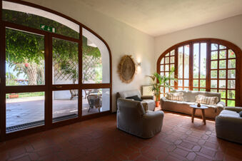 A seating area at B&B HOTEL Ischia San Nicola with beige armchairs and sofa, wooden table, large windows, and outdoor greenery visible