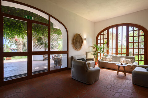 A seating area at B&B HOTEL Ischia San Nicola with beige armchairs and sofa, wooden table, large windows, and outdoor greenery visible