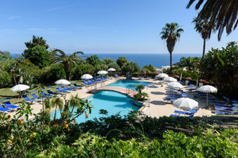 Swimming pool with blue sun loungers, white umbrellas, palm trees, and ocean view at B&B HOTEL Ischia San Nicola