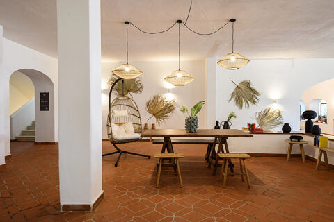 Open lobby area of B&B HOTEL Ischia San Nicola with terracotta floor tiles, wooden table, benches, hanging rattan lights, and hanging chair