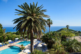 B&B HOTEL Ischia San Nicola outdoor pool area with palm trees, blue lounge chairs, white umbrellas, and a view of the sea and hills