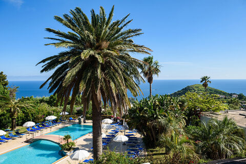 B&B HOTEL Ischia San Nicola outdoor pool area with palm trees, blue lounge chairs, white umbrellas, and a view of the sea and hills