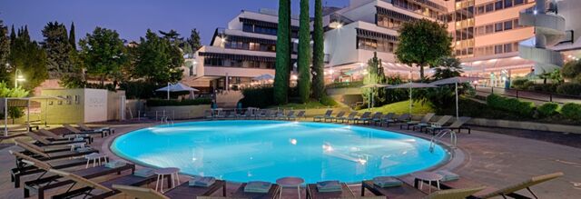 Outdoor round swimming pool surrounded by lounge chairs and umbrellas at Hotel NH Roma Villa Carpegna during evening time